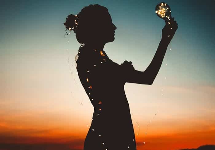 Artistic silhouette of a woman wrapped in fairy lights during a vibrant sunset.